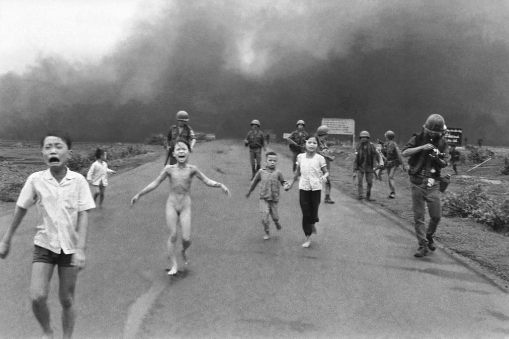 Five children are running down the road, crying. One of the girls is naked. Behind the children there are seven soldiers. The sky is dark and full of smoke. Black and white picture from the Vietnam war. Photo.  
