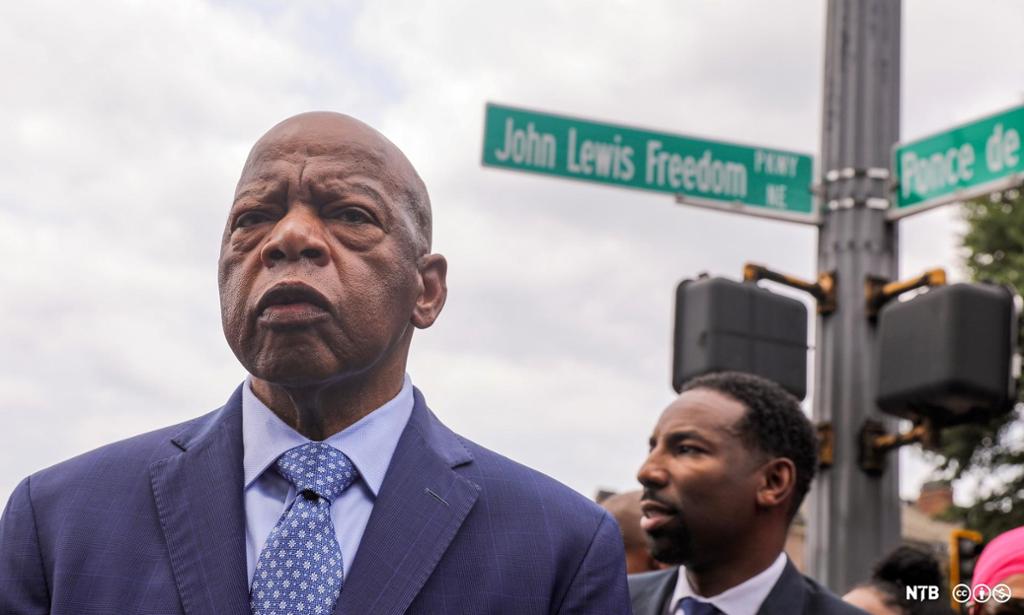 Photo: We see John Lewis, he is an older Black man. He is bald. He is wearing a blue suit with a tie. Behind him we see a younger man. There is a street sign behind them. The sign says John Lewis Freedom. 