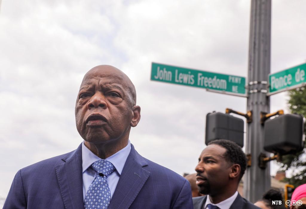 Photo: We see John Lewis, he is an older Black man. He is bald. He is wearing a blue suit with a tie. Behind him we see a younger man. There is a street sign behind them. The sign says John Lewis Freedom.
