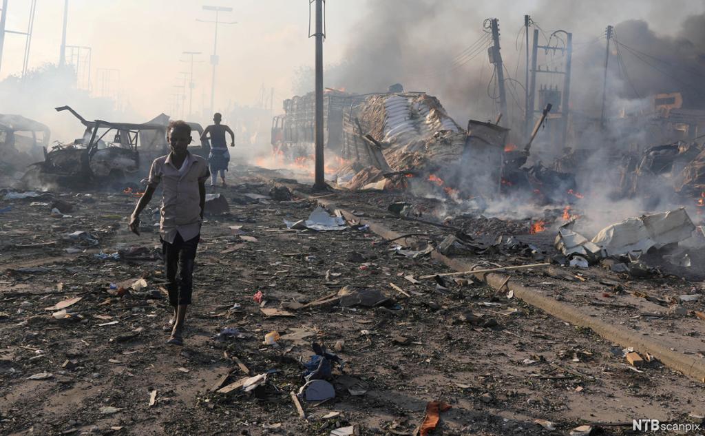 Explosion in Mogadishu, Somalia. Burning cars and debris in the street, with a few civilians leaving the area. Photo.