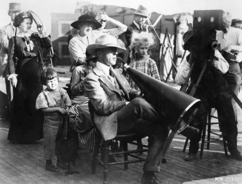 En mann sitter på en stol med en ropert ved siden av en filmfotograf og omgitt av tilskuere. Foto.