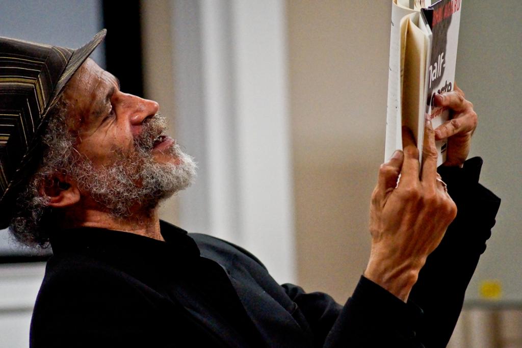 The British-Caribbean poet John Agard is reading from one of his books. He's a coloured man with a grey beard. Photo.