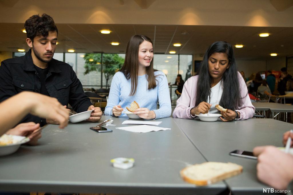 Elever spiser sammen i kantina. De spiser fra tallerkener og har brødskiver i handa. Et par mobiltelefoner ligger på bordet. Foto. 