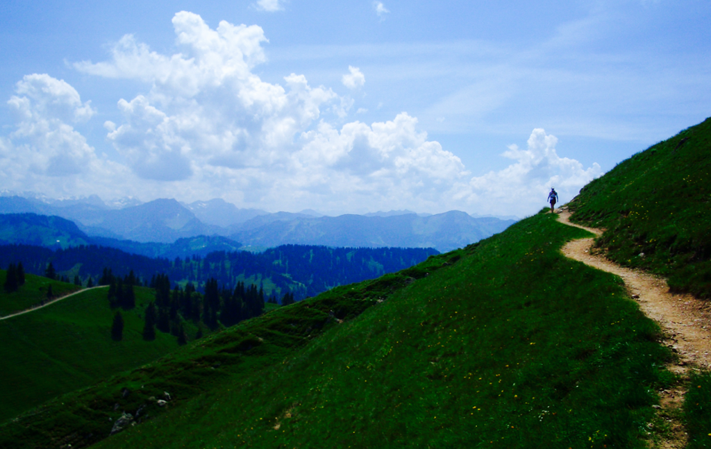 Sti i grønt alpelandskap. En person er på fjelltur. Nydelig utsikt mot en blå horisont. Foto.