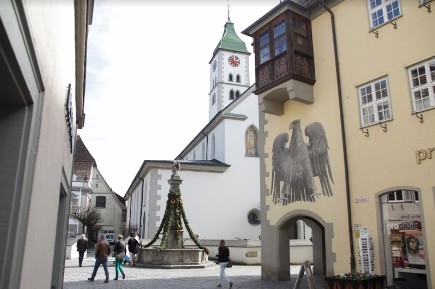 Wangen i Tyskland: torg med brønn som er pynta med grangirlander. Ved torget ei kirke og en bygning med et stort våpenskjold og et gammelt karnappvindu på veggen. Foto.