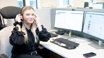 Jente i sort tøy sitter med et headset ved arbeidspulten sin i et åpent kontormiljø. Foto.
