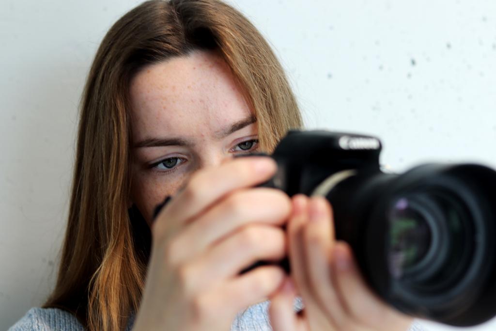 Nærbilde av en elev som holder et speilreflekskamera som hun ser på. Kameraet er i forgrunnen. Fokuset ligger på ansiktet til jenta. Foto.