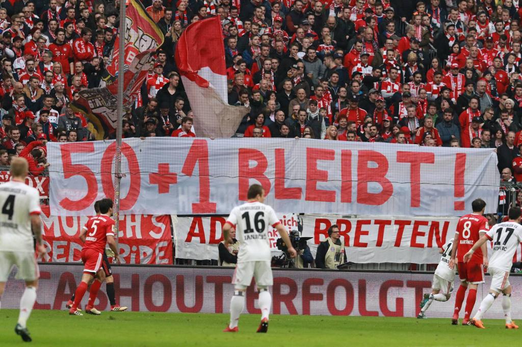 Fotballkamp med spillere i forgrunnen og publikum i bakgrunnen. På tribunen holdes det opp et banner med påskriften "50+1 BLEIBT!". Foto.