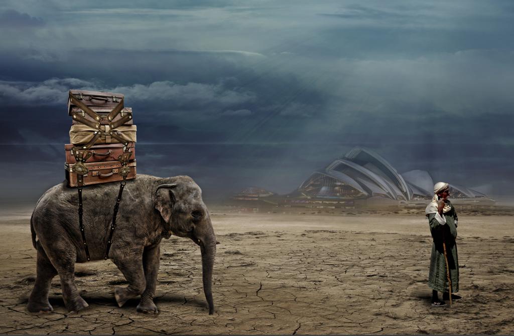 Photo art: Desert landscape. An elephant has many suitcases piled on its back. A man dressed in traditional Middle Eastern clothes stands looking out of the picture to the right. In the background we see a building that looks like the Opera House in Sydney, Australia. 