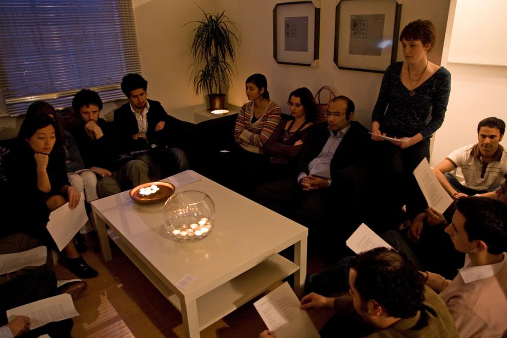 Mange personer er samlet i ei stue rundt et bord. Noen holder trykte hefter i hendene. En står opp og leser noe fra et hefte. Foto.