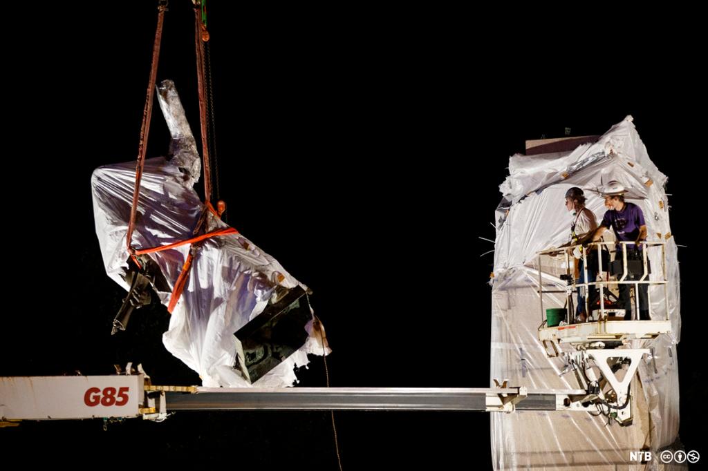 Sculpture wrapped in plastic is hoisted in the air by a crane. Two men are on the crane. 