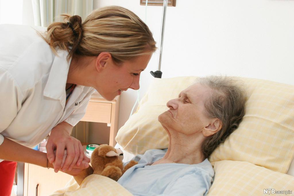 En kvinnelig helsefagarbeider holder hånda til en gammel kvinne som ligger i ei seng. Hun bøyer seg nært kvinnens ansikt og smiler til henne. Foto.