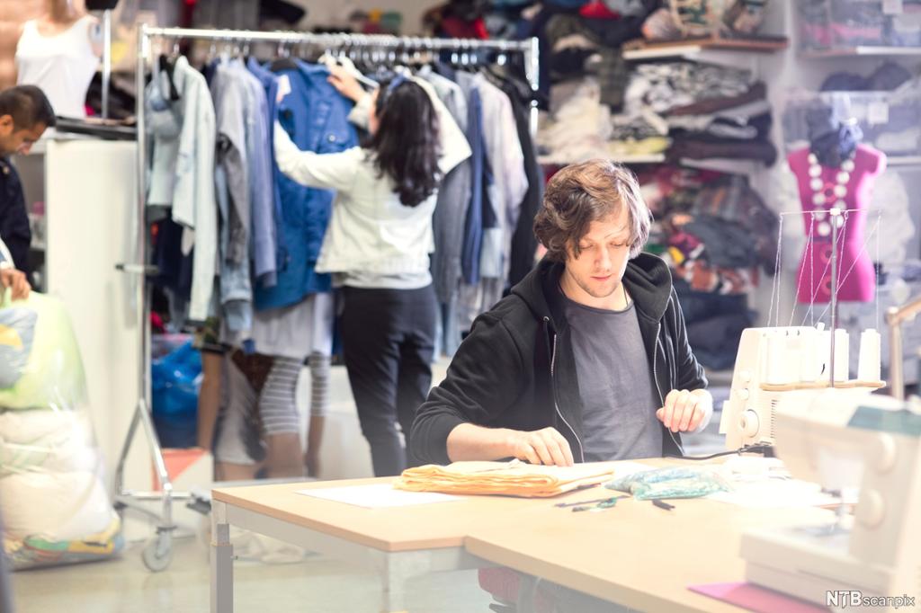 Ung mann sitter ved symaskin med ung kvinne som leter etter klær på et klesstativ i bakgrunnen. Foto.