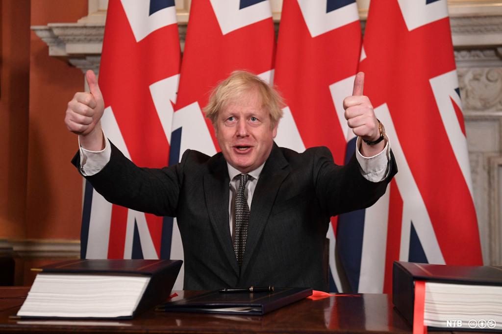 Photo: Boris Johnson. We see a blond man in a dark suit and tie. He is giving two thumbs up. Behind him is a row of British flags. 