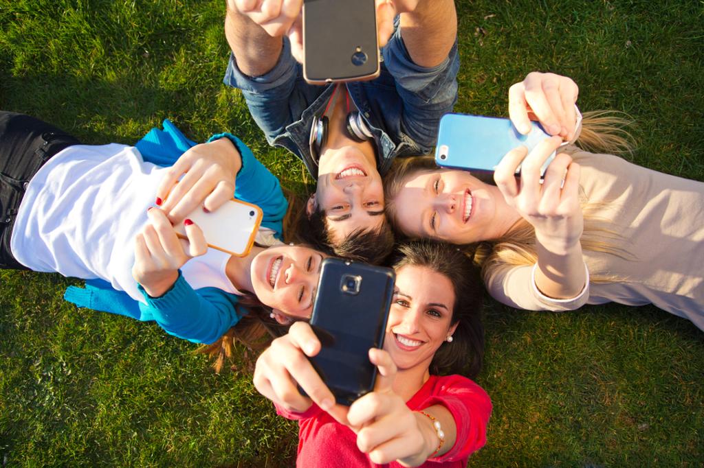 Fire ungdommer ligger på ei grønn plen med hodene mot hverandre. Alle holder opp hver sin mobiltelefon og tar en selfie mens de smiler. Foto.