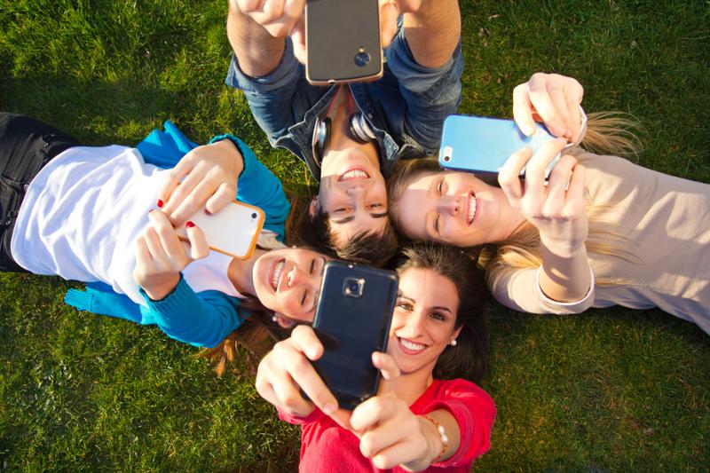 Fire smilende ungdommer ligger på ei mark og tar en selfie. Foto.