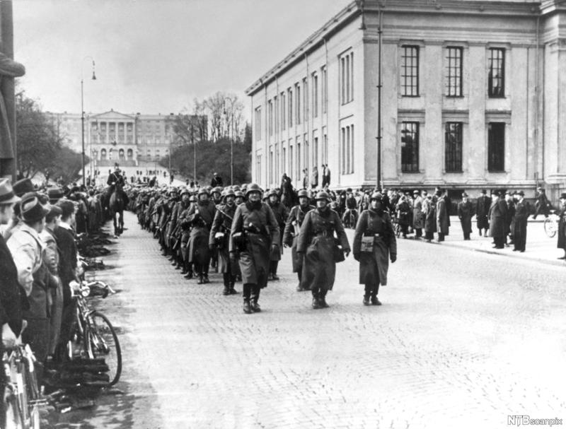 Svart-hvitt fotografi av tyske tropper i uniformer med lange frakker. De marsjerer på Karl Johans gate ved universitetsplassen. Slottet ligger bakerst i bildet. Bildet er 9. april 1940. 