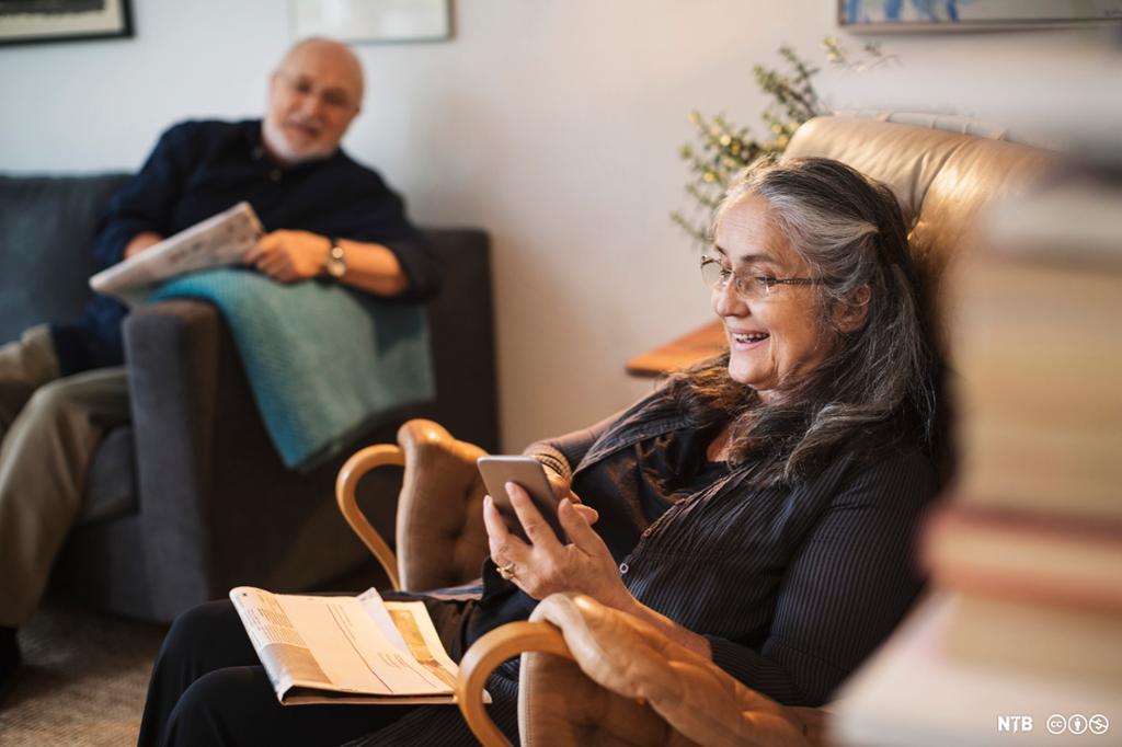 Eldre dame med avis i fanget ser på mobilen. Hun smiler. I bakgrunnen sitter en eldre mann med avis i en sofa og ser bort på kvinnen. Foto.