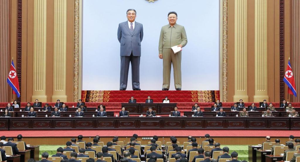 To enorme, naturtro  statuer står på en scene bakerst i en sal. Foran dem sitter  to-tre rekker på et opphøyd podium. I salen, vendt mot dem, sitter mange på rekke og rad. Nordkoreanske flagg står på hver side. Foto.