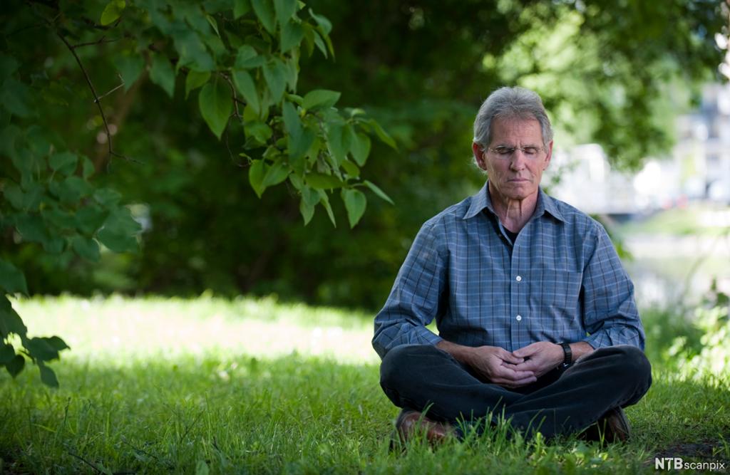 Mann mediterer utandørs. Han sit i grønt gras med lukka auge og beina i kross. Foto.