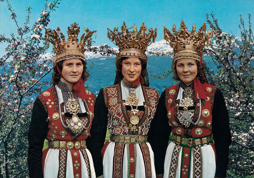 Tre unge kvinner i tradisjonelle folkedrakter, med broderi, forkle og søljer. Alle tre har store brudekroner. I bakgrunnen er det blomstrande frukttre og snøkledde fjelltoppar. Foto.