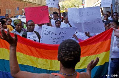 Photo: We see protesters with cardboard signs behind a rainbow flag. 