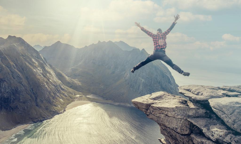 En person poserer på en fjelltopp. Bildet er tatt idet hen gjør et hopp opp i lufta. Det er vakker utsikt til fjell og hav og fine og stille værforhold. Fotografi.