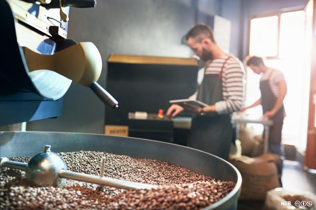 I forgrunnen males kaffebønner i en stor maskin. I bakgrunnen står to menn med lange forklær og jobber med andre deler av kaffeprosessen. Foto.