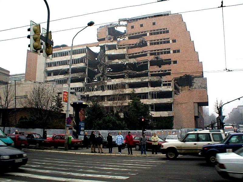 Øydelagd bygning etter bombing. Det er folk og bilar i forkant av bygninga. Foto.