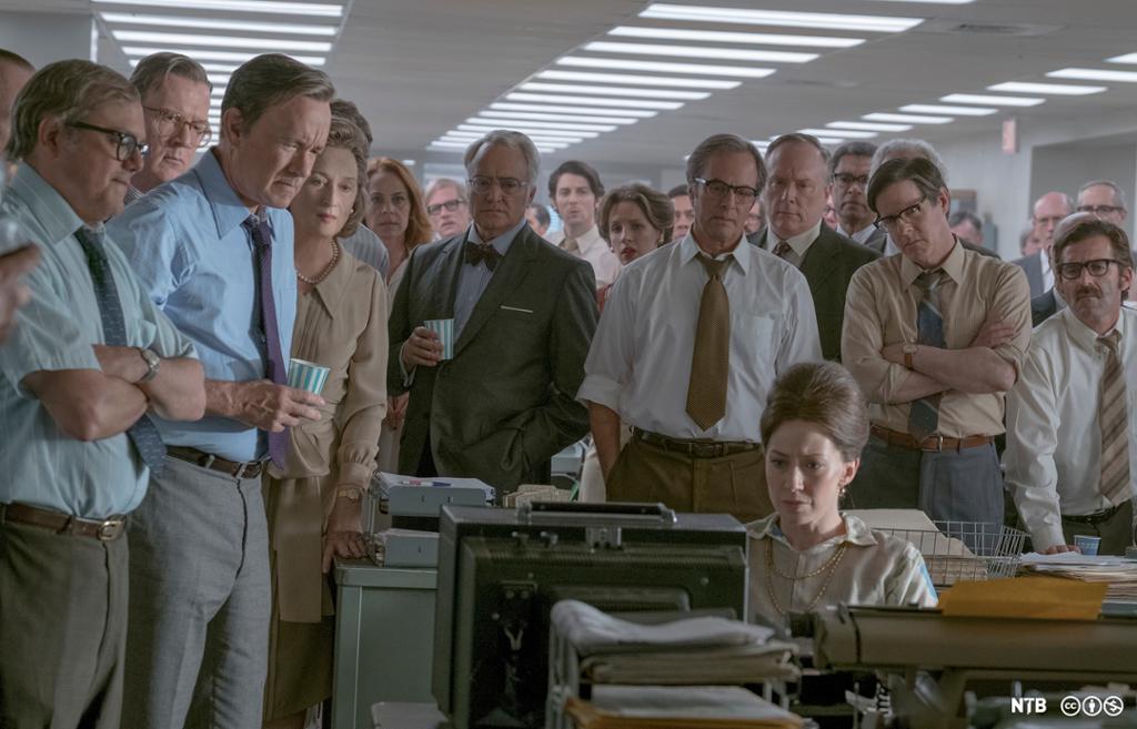 Ei gruppe menn og noen få kvinner står i ring rundt en kvinne som studerer dokumenter i en avisredaksjon. Foto.